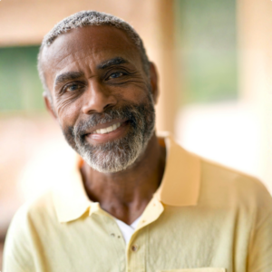 An older man with short gray hair and a beard smiles warmly. He is wearing a light yellow collared shirt and is standing in a softly blurred outdoor setting.