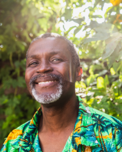 A smiling man with short hair and a gray beard wears a tropical shirt while standing outside in bright sunlight, surrounded by green foliage.
