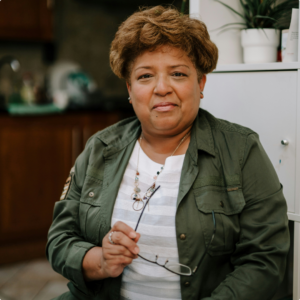 A woman with short curly hair sits indoors, holding glasses in one hand and smiling gently. She wears a green jacket over a white top and a necklace, with shelves and a plant in the background.