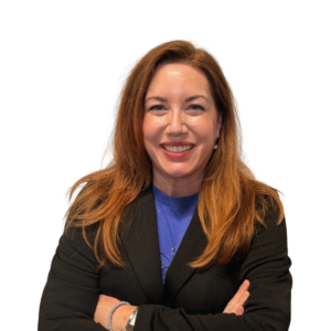 A woman with long auburn hair wearing a black blazer and blue shirt stands against a white background, smiling with arms crossed.