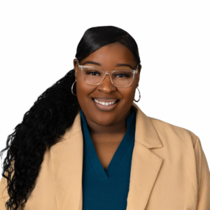 A woman with long curly hair in a ponytail, wearing clear glasses, hoop earrings, a beige blazer, and a teal top, smiles at the camera against a plain white background.