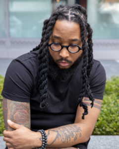A man with long braided hair, round glasses, and tattoos sits outdoors, looking down with his arms crossed, wearing a black t-shirt and beaded bracelets.