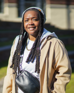 A person with long dreadlocks, wearing a beige coat, gray beanie, white shirt, and a crossbody bag, stands outdoors in sunlight, smiling.