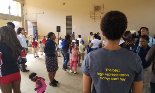 A group of people, including children and adults, gather indoors for an event. One person in the foreground wears a shirt reading "The best legal representation money can't buy." Some children are playing while others stand and talk.