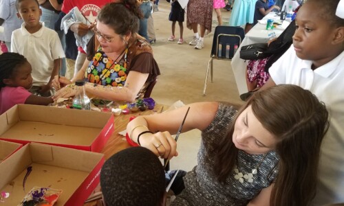 A woman paints a boy’s face at a busy indoor community event. Children and adults are gathered around tables with art supplies, socializing and participating in activities.