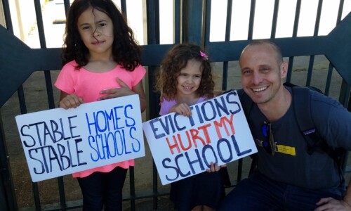 Two young girls hold signs reading "Stable Homes Stable Schools" and "Evictions Hurt My School" while standing next to a smiling man crouched beside them. They are outdoors in front of a metal gate.