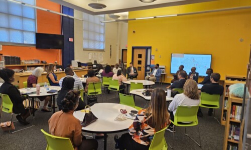 A group of adults sits in a classroom with bright yellow and blue walls, listening to speakers at the front near a large screen. Most people are seated at round tables, facing forward, and the room is well-lit.