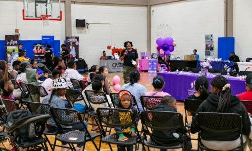 A community event in a gymnasium with people seated, children and adults listening to a woman speaking. Tables with purple decorations and balloons are set up, and informational booths line the gym walls.
