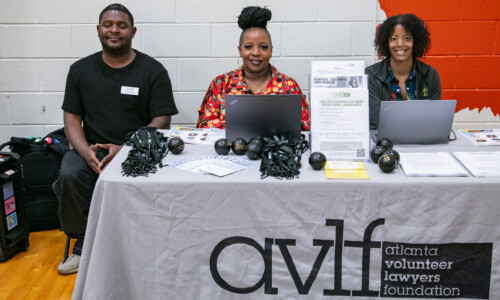 Three people sit behind a table covered with an AVLF (Atlanta Volunteer Lawyers Foundation) tablecloth, with laptops, flyers, and stress balls, at an indoor event in front of a white and orange wall.