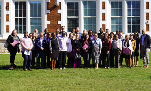 A large group of people posing and smiling outside in front of a building with many windows. Some are holding folders and wearing name tags, and the group is standing on green grass in sunlight.