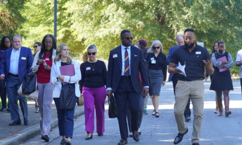 A group of professionally dressed adults, some holding folders, walk together outside on a sunny day along a tree-lined street, engaging in conversation.