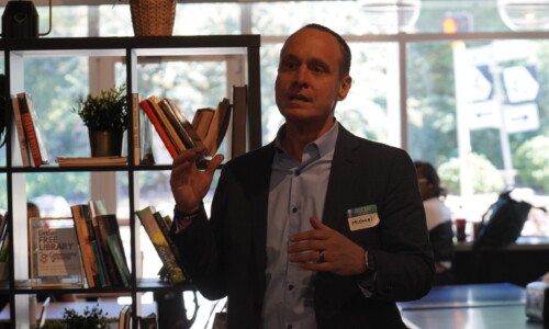 A man in a suit with a name tag gestures while speaking in front of a bookshelf at an indoor event, with large windows and people visible in the background.