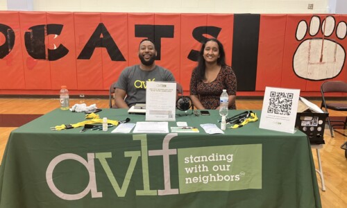 Two people sit behind a green table with the AVLF logo and “standing with our neighbors” text. The table has flyers, pens, and a QR code. Behind them is a gym wall with "CATS" and a paw print.