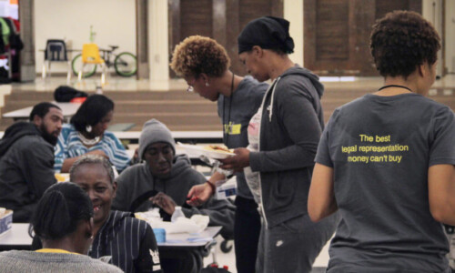 People eat and talk at tables in a large cafeteria. Three women in gray shirts serve food; one shirt reads, “The best legal representation money can’t buy.” Some people appear to be volunteers helping others.