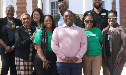 A group of nine adults pose and smile outdoors in front of a brick building. Some wear business attire while others wear green t-shirts. The group stands closely together in bright sunlight.
